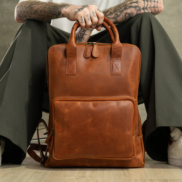 A person with tattooed arms holds the Bolton Leather Backpack – Tan Brown by Vintage Leather Sydney while sitting on the floor; only their lower body, hands, and part of their sneakers are visible.