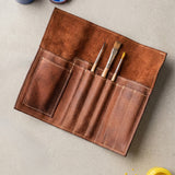 The Leather Artist Roll Zip by Vintage Leather Sydney, a brown leather pouch, neatly holds four paintbrushes and is shown on a light surface near yellow and blue paint containers.